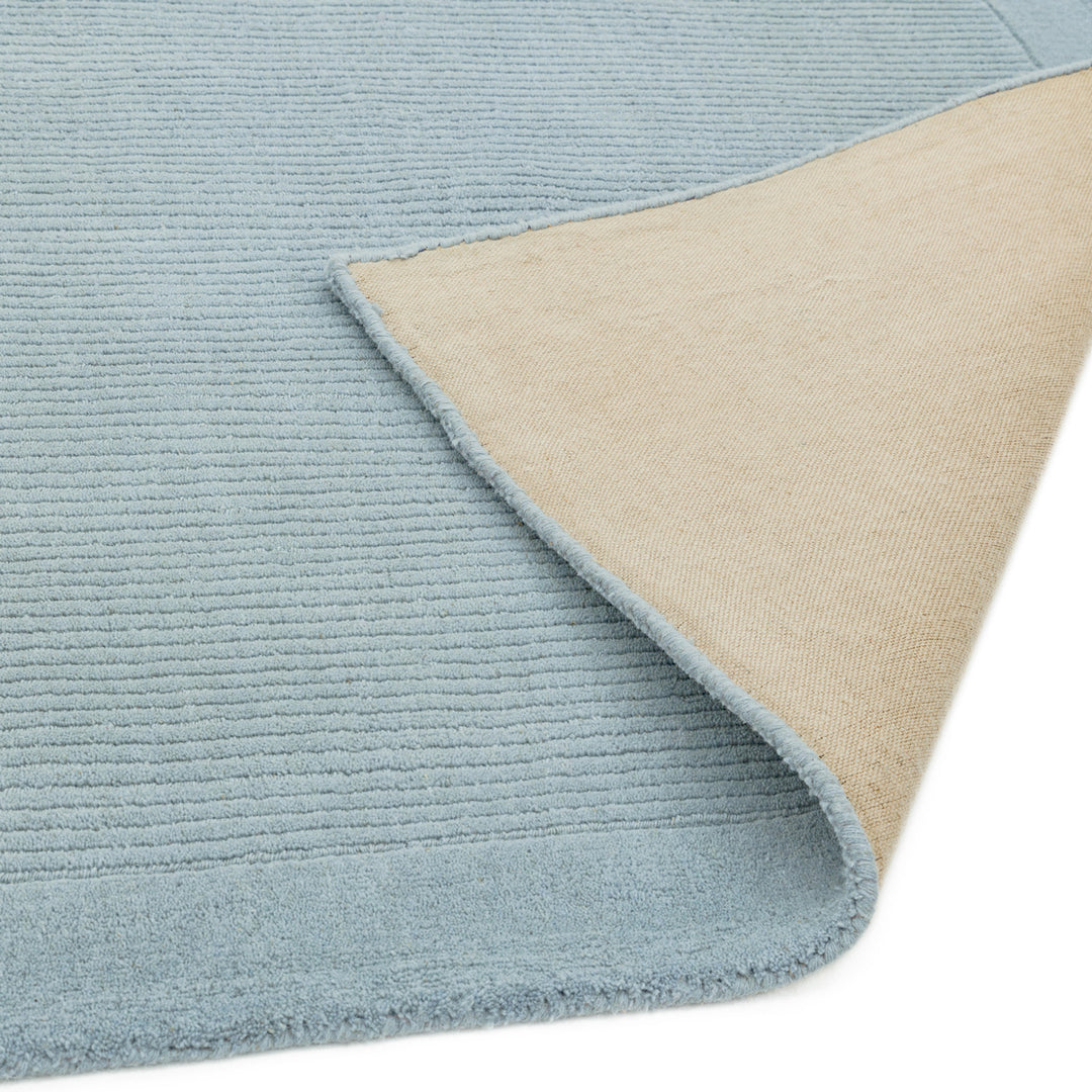 澄み渡った空を想わせる淡いベビーブルー色に、リブとボーダーがスタイリッシュ感を添える、サステナブルな天然素材ウール(羊毛)100%製RUGHAUSのふんわりシンプル手織りラグの素材感と裏地がわかるクロースアップ
